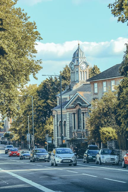 A busy street scene in Highbury featuring a row of parked cars along the pavement, with various models and colours of vehicles lined up in front of a historic church building. The church has a prominent clock tower with a domed spire, large arched windows, and decorative brickwork, surrounded by lush green trees that partially obscure its facade. The street is illuminated by tall lampposts, and the sky above is bright with a few scattered clouds. This image illustrates the typical environment encountered during house relocations or moving services carried out by Man With a Van Highbury, highlighting the urban setting and transportation elements involved in furniture transport and home moving processes.