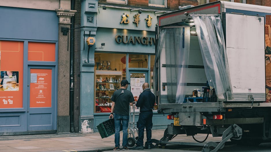 Two men are engaged in a furniture transport and home relocation process outside a retail establishment named 'Guanghe'. One individual, wearing a black shirt and jeans, is holding a wheeled dolly, while the other, dressed in a dark jacket and pants, appears to be assisting or supervising. They are standing on a paved sidewalk adjacent to a loading area where a large, grey delivery truck with an open, semi-transparent plastic curtain on the side is parked. The truck's cargo area contains packaging materials and possibly furniture or boxes ready for transportation. The shop window behind them displays various items and has signs with Chinese characters and English text. The building facade is classical with architectural details, and the overall environment suggests a street scene during daylight, with ambient lighting indicating natural daylight. Man With a Van Highbury is known for such removals and transport services, supporting efficient packing and furniture moving logistics.