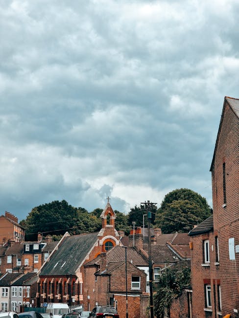 A view of a residential street in Highbury with red brick houses and a small church with a peaked roof and a cross on top, framed by trees and partly cloudy sky. In the foreground, there are several cardboard boxes and a large plastic-wrapped package placed on the pavement, indicating packing for a home relocation. A black van with an open rear door is parked nearby, featuring visible moving blankets, straps, and equipment used during the furniture transport and loading process. The scene captures the behind-the-scenes details of a professional house removal, with furniture and boxes prepared for loading into the vehicle, reflecting the logistics involved in local removals for the Highbury area, as offered by Man With a Van Highbury.