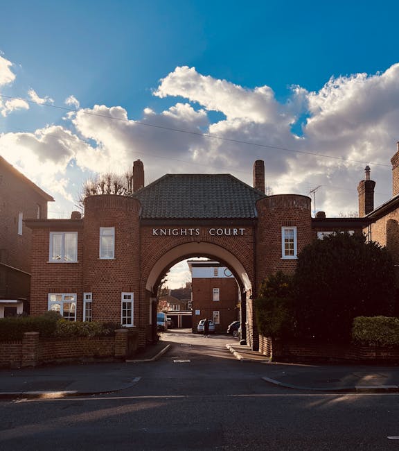 A brick-built residential building with an arched entrance labeled 'Knights Court' serves as the gateway to a driveway where multiple cardboard boxes, wrapped furniture, and packing materials are being loaded into a white van parked just outside the archway. The area is shaded by nearby trees and bushes, with some items apparently being carried or positioned on trolleys by movers. The background features a partly cloudy sky with sunlight illuminating parts of the scene, indicating daylight hours. This image illustrates the moving process at a property in Highbury, captured by Man With a Van Highbury during a home relocation involving furniture and packing logistics at the Knights Court estate.