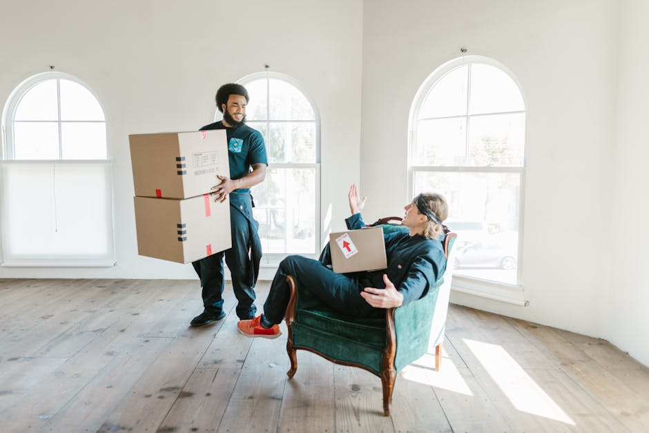 A male mover from Man With a Van Highbury, dressed in casual dark clothing, is inside a spacious, well-lit room with white walls and three large arched windows. He is holding two cardboard boxes stacked on top of each other, secured with red packing tape. A woman, seated in a vintage green armchair positioned near the windows, is lifting her hand as if to signal or gesture. She is also holding a cardboard box with a white sticker and red arrow indicating fragile or priority items. The room has a light wooden floor, and sunlight streams through the windows, illuminating the scene with natural light. The image captures the process of home relocation, with furniture and packing materials visible, reflecting a furniture transport and packing and moving activity typical of house removals carried out by Man With a Van Highbury.