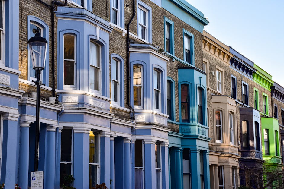 Exterior view of a row of Victorian-style terraced houses in Highbury with colorful facades in shades of blue, beige, brown, and green. The buildings feature large bay windows, decorative moldings, and brickwork, with some windows reflecting the sky and surroundings. A black street lamp stands on the pavement in front of the houses. The scene captures a house removal process, where [COMPANY_NAME], a professional removals service, is likely involved in loading and transporting household furniture and boxes from inside the property through the doorway, possibly using equipment such as trolleys or blankets visible in the context of a home relocation or furniture transport. Natural daylight illuminates the scene, emphasizing the architectural details and preparing for the next stage of packing and moving activities associated with house removals in the Highbury area.