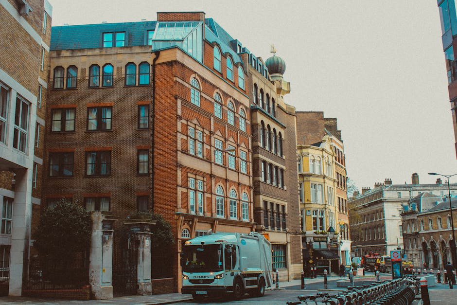 A street scene in Highbury showing a small white van parked adjacent to a sidewalk with a row of modern and historic multi-storey buildings in the background. The buildings feature a mix of brick and stone facades with large windows, some with arched tops, and decorative architectural details. The van is situated near the entrance of an alleyway or driveway, indicating a home relocation or furniture transport process. Nearby are bicycles secured along the street, and the pavement appears clean and orderly. The scene is captured during daytime with overcast lighting, providing clarity of the surroundings. This image exemplifies the typical environment encountered during professional removals and house moving services provided by [COMPANY_NAME], with visible elements related to packing, loading, and transport of household items in an urban setting effectively illustrating the logistical aspect of house removals in Highbury.