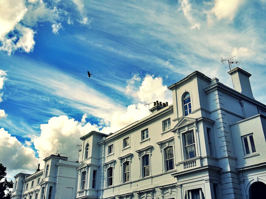 Exterior view of a row of Victorian-style terraced houses in Highbury with colorful facades in shades of blue, beige, brown, and green. The buildings feature large bay windows, decorative moldings, and brickwork, with some windows reflecting the sky and surroundings. A black street lamp stands on the pavement in front of the houses. The scene captures a house removal process, where [COMPANY_NAME], a professional removals service, is likely involved in loading and transporting household furniture and boxes from inside the property through the doorway, possibly using equipment such as trolleys or blankets visible in the context of a home relocation or furniture transport. Natural daylight illuminates the scene, emphasizing the architectural details and preparing for the next stage of packing and moving activities associated with house removals in the Highbury area.