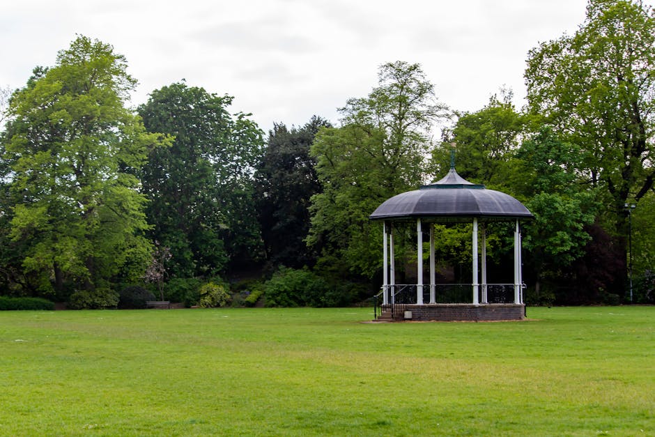 A spacious park area features a well-maintained green lawn with evenly cut grass, surrounded by tall, leafy trees with varying shades of green foliage. In the center-right of the image, there is a small, circular bandstand or gazebo with a dark-colored, domed roof supported by white columns, elevated on a low brick platform. The bandstand is enclosed by a metal railing painted black, and there are steps leading up to its entrance. The background includes a dense cluster of trees creating a natural backdrop for the open space. The sky above is overcast with grey clouds, providing diffuse lighting across the scene. This setting illustrates a typical outdoor location suitable for communal gatherings, and it aligns with the kinds of environments where house removals and furniture transport services may operate, as provided by Man With a Van Highbury.