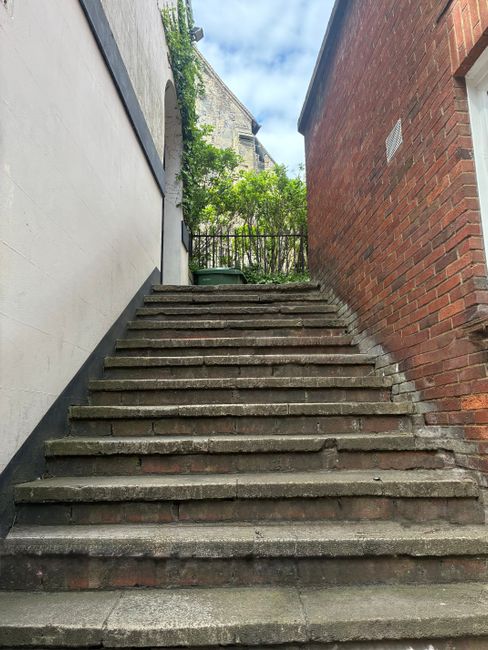 A straight staircase inside a building with white walls and a large window on the right side allowing natural light to illuminate the space. The staircase has red-brown tiled steps with white risers, 