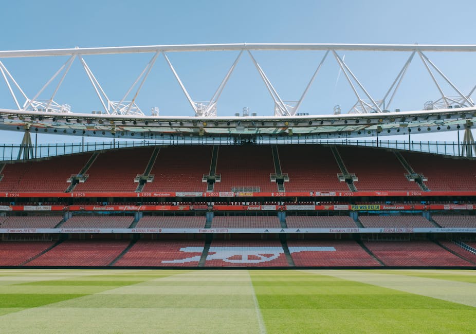 An exterior view of Emirates Stadium with clear blue sky overhead, featuring red seating tiers, some with advertisements and banners, and a green grass pitch in the foreground. The stadium's large white roof structure, supported by a network of trusses, extends over the seating area. Inside the stadium, no furniture or moving objects are visible, as the image captures a wide-angle shot of the sports venue, illustrating its architecture and community presence. Although the stadium is not directly related to house removals or relocation services, the image provides context about the location near Highbury, where [COMPANY_NAME] offers house removals and moving logistics. The photo emphasizes the structure and environment of the stadium during daylight, with no ongoing moving or loading activities present.