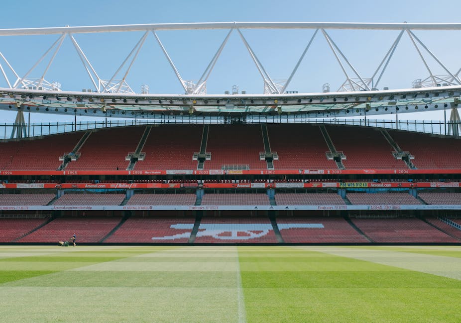 Inside the Emirates Stadium, the image shows the view from the pitch level towards the empty red seating areas, with the large white overhang roof structure visible above. The stadium's seating sections are arranged in tiers, with some sections displaying sponsor banners and team logos, such as Arsenal. The bright green grass of the football field is in the foreground, with a worker using a lawn mower or maintenance equipment on the turf. Natural daylight illuminates the scene, highlighting the clean, modern architecture of the stadium's roof and seating areas. This image connects to the theme of house removals and relocation services by illustrating a large, spacious environment associated with high-profile venues, echoing the logistics of packing, transportation, and arrangement involved in professional moving services. Man With a Van Highbury, experienced in removals, might arrange transport or storage for items destined for such venues or similar settings in the context of home or furniture move processes.