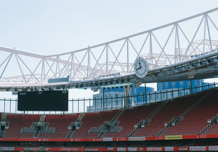 An exterior view of Emirates Stadium with clear blue sky overhead, featuring red seating tiers, some with advertisements and banners, and a green grass pitch in the foreground. The stadium's large white roof structure, supported by a network of trusses, extends over the seating area. Inside the stadium, no furniture or moving objects are visible, as the image captures a wide-angle shot of the sports venue, illustrating its architecture and community presence. Although the stadium is not directly related to house removals or relocation services, the image provides context about the location near Highbury, where [COMPANY_NAME] offers house removals and moving logistics. The photo emphasizes the structure and environment of the stadium during daylight, with no ongoing moving or loading activities present.