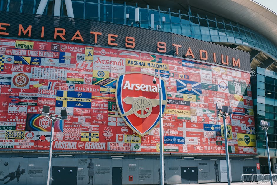 Exterior view of the Emirates Stadium showing a large, colorful mosaic featuring various football club logos, flags, and supporters' banners, prominently displaying the Arsenal Football Club crest at the center. The stadium's name, 'Emirates Stadium,' is visible at the top of the building, which features a modern glass and steel façade. In the foreground, several tall floodlights and security cameras are mounted on poles, illuminating the area around the stadium entrance. The scene is captured during daytime with clear lighting, highlighting the detailed mosaic artwork on the stadium's exterior, which celebrates football supporters and clubs from around the world. This detailed depiction emphasizes the large scale of the stadium, suitable for team events and home matches, and reflects the context of a major sports venue, relevant to event logistics and fan transportation services provided by Man With a Van Highbury.