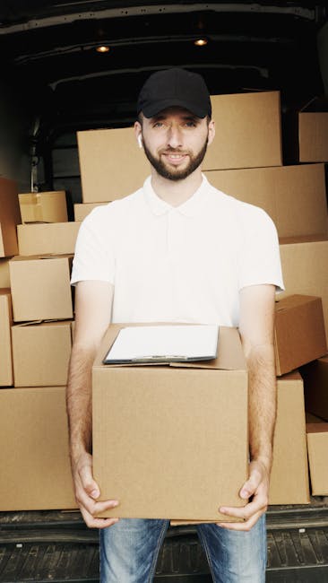 A man wearing a black cap and a white t-shirt is standing inside the open back of a van, holding a cardboard box in front of him. The interior of the van is filled with neatly stacked cardboard boxes of various sizes, taped and ready for transport, likely part of a home relocation or moving process. The man has wireless earbuds in his ears and is outdoors on a paved surface, with natural daylight illuminating the scene. The background shows the dark interior of the van, emphasizing the loading and packing activities typical of furniture transport and moving services provided by Man With a Van Highbury. The image captures the loading process involved in packing and organizing household items for a quick and efficient move.