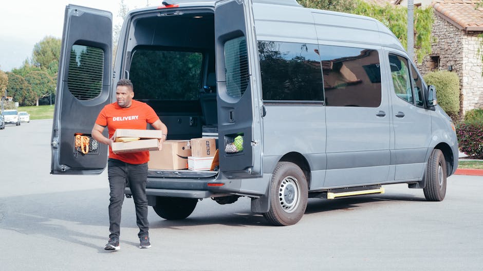 A man wearing a red T-shirt with the word 'DELIVERY' printed on it, standing on a paved surface outside a residential area, is holding two cardboard boxes filled with packing materials and smaller boxes. Behind him, a grey panel van with its rear doors open reveals a variety of packed moving boxes, some wrapped in plastic and others unwrapped, as well as a few envelopes and wrapping materials stored inside. The van is positioned on a driveway near a house with a brick exterior and tiled roof, with greenery and trees visible in the background. The scene depicts a home relocation or furniture transport process, with a focus on loading or unloading activities, supported by the professional moving service provided by Man With a Van Highbury.