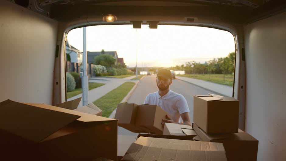 A man wearing a black cap and a white t-shirt is standing inside the open back of a van, holding a cardboard box in front of him. The interior of the van is filled with neatly stacked cardboard boxes of various sizes, taped and ready for transport, likely part of a home relocation or moving process. The man has wireless earbuds in his ears and is outdoors on a paved surface, with natural daylight illuminating the scene. The background shows the dark interior of the van, emphasizing the loading and packing activities typical of furniture transport and moving services provided by Man With a Van Highbury. The image captures the loading process involved in packing and organizing household items for a quick and efficient move.